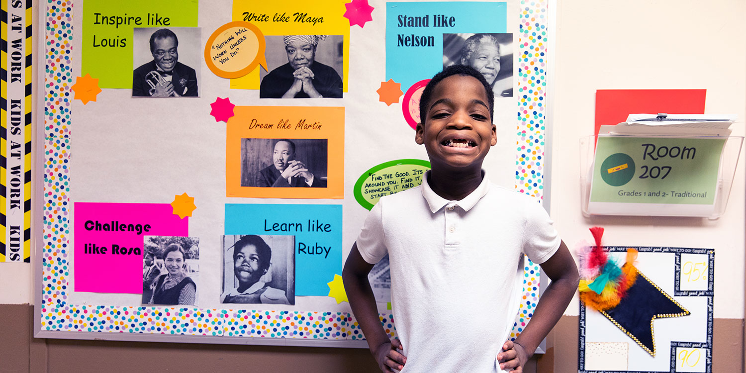 Elementary student standing in front of decorated bulletin board with big smile.